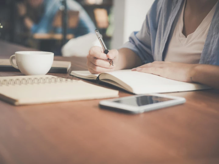 Woman Writing on a Notebook Beside Teacup and Tablet Computer