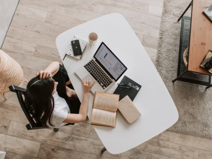 From above of young woman with long dark hair in casual clothes working at table and browsing netbook while sitting in modern workplace and touching hair