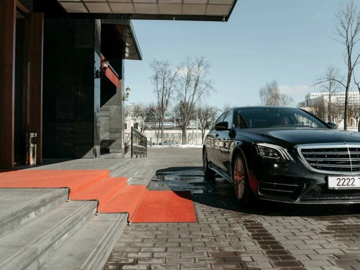 A luxury black sedan parked on a red carpet driveway outside a modern building.