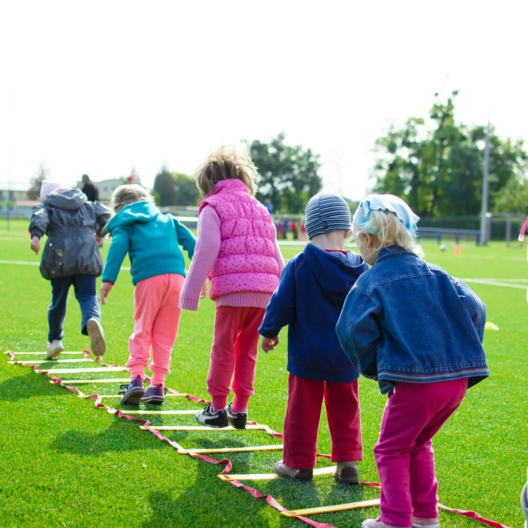 children enjoy an outdoor activity on a grassy field stepping over a ladder